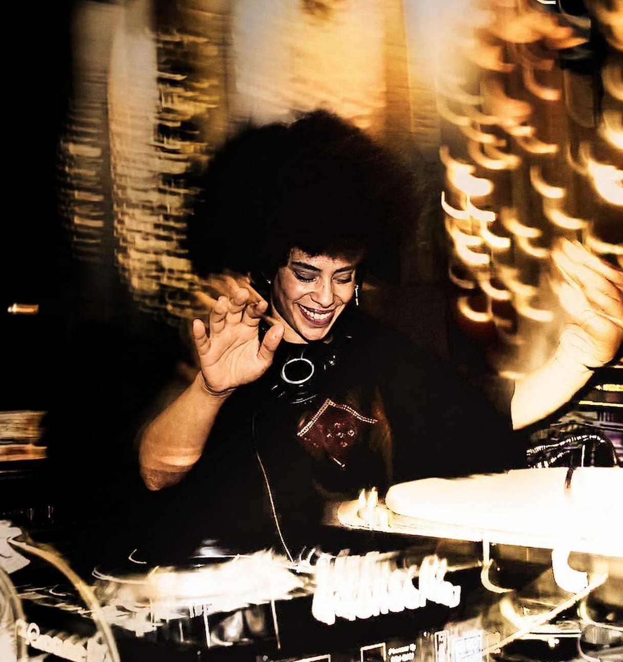 Allynx behind the decks, hands raised, warm light trails around her.