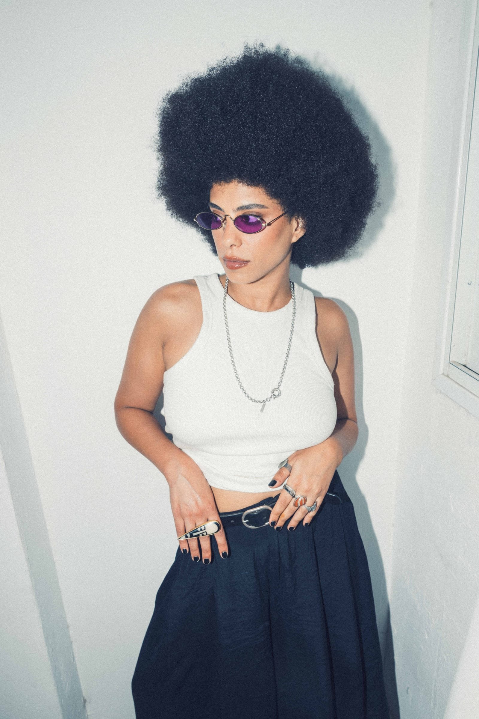 Allynx standing against a white wall in a white top and dark trousers, wearing tinted round sunglasses.