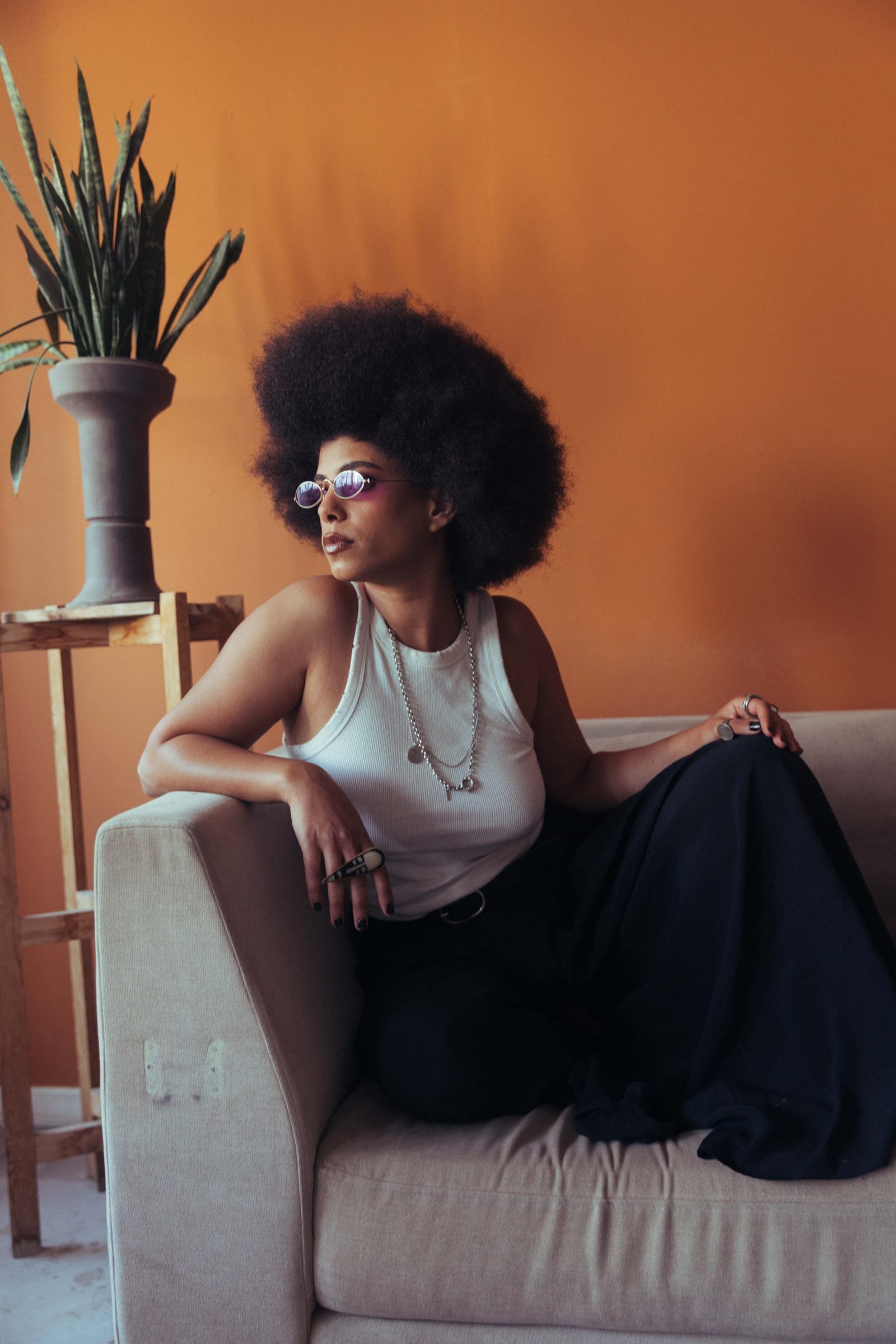 Allynx seated in front of a warm orange wall, wearing tinted round sunglasses.